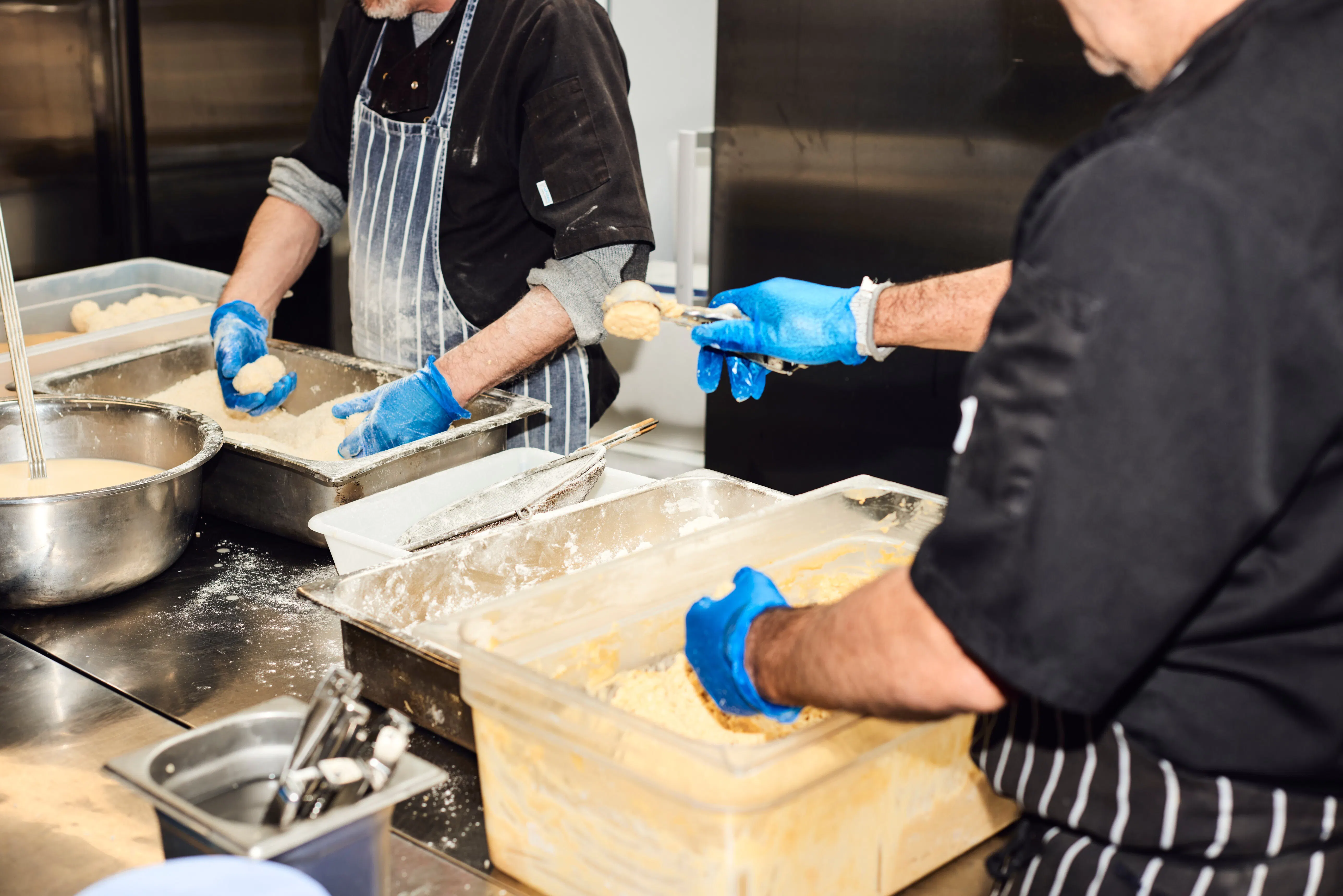Two chefs in blue gloves breading portions of food in gastronorm trays at a floured prep bench